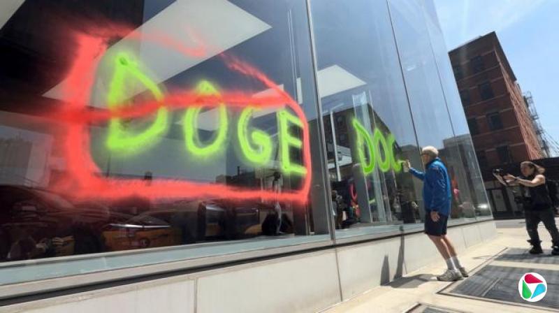 特斯拉的窗户被喷涂着“DOGE”字样,并用红色圆圈和斜线划掉,旁边站着一名男子,开始喷涂另一句口号。