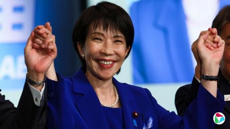 高市早苗笑容灿烂。她举起双手庆祝,与镜头外的另外两人的双手交握??。她穿着宝蓝色套装,戴着珍珠耳环和珍珠项鍊。