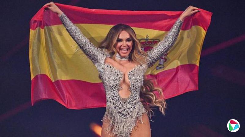 西班牙歌手梅洛迪(Melody)身穿银色舞台服装,身后举着西班牙国旗,面带微笑。