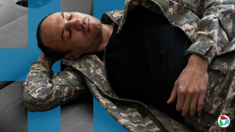 一名身穿迷彩外套和裤子的士兵躺在沙发上,一只手托着头。他双眼紧闭,仿佛已经或即将入睡。