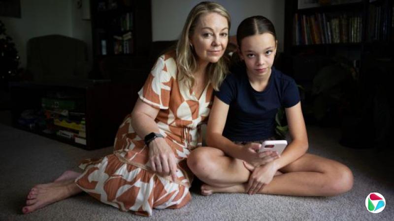 一名身穿棕白色洋装的女子坐在铺有地毯的地板上,身旁是她的青少女女儿,手中拿着一支粉红色手机